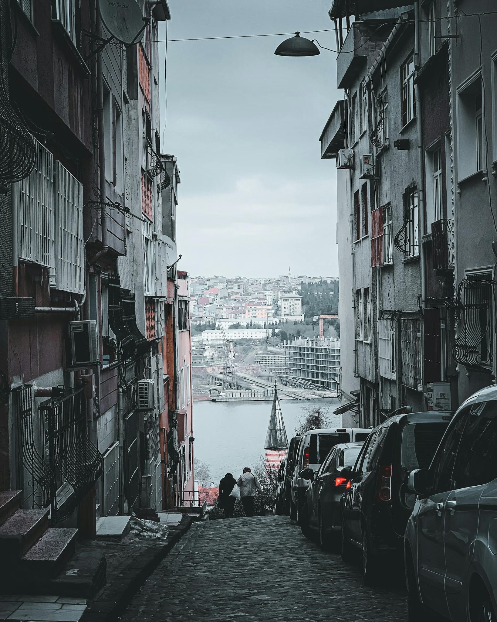 Narrow urban alley with cobblestone street leading to a scenic waterfront view, flanked by buildings.